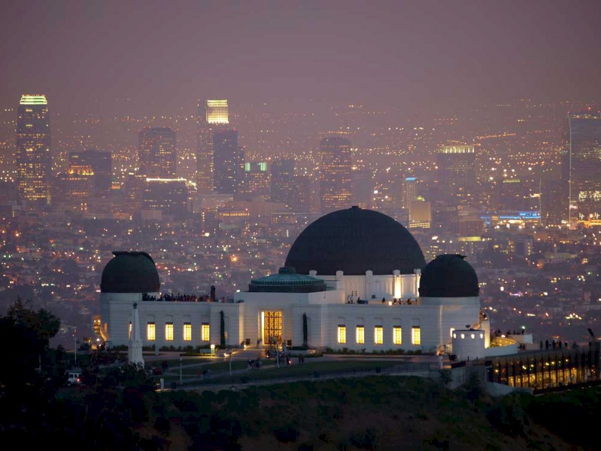 A domed observatory building is illuminated at night, with a city skyline full of lights and tall buildings in the background.