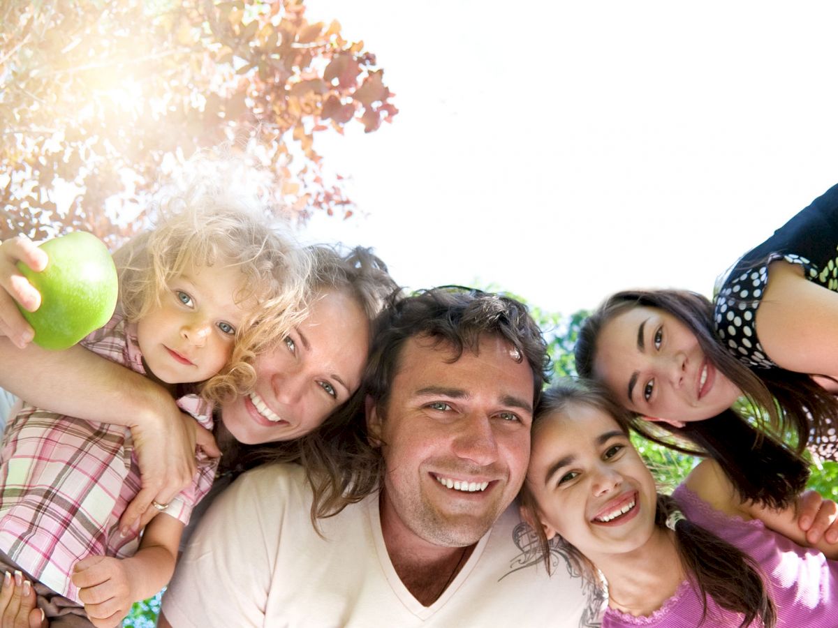 A group of five people, including children and adults, smiling and posing together outdoors with greenery in the background.