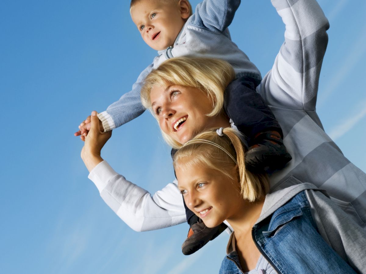 A cheerful trio enjoys a sunny day; one child is on an adult's shoulders, while another stands beside them, all smiling and looking up.