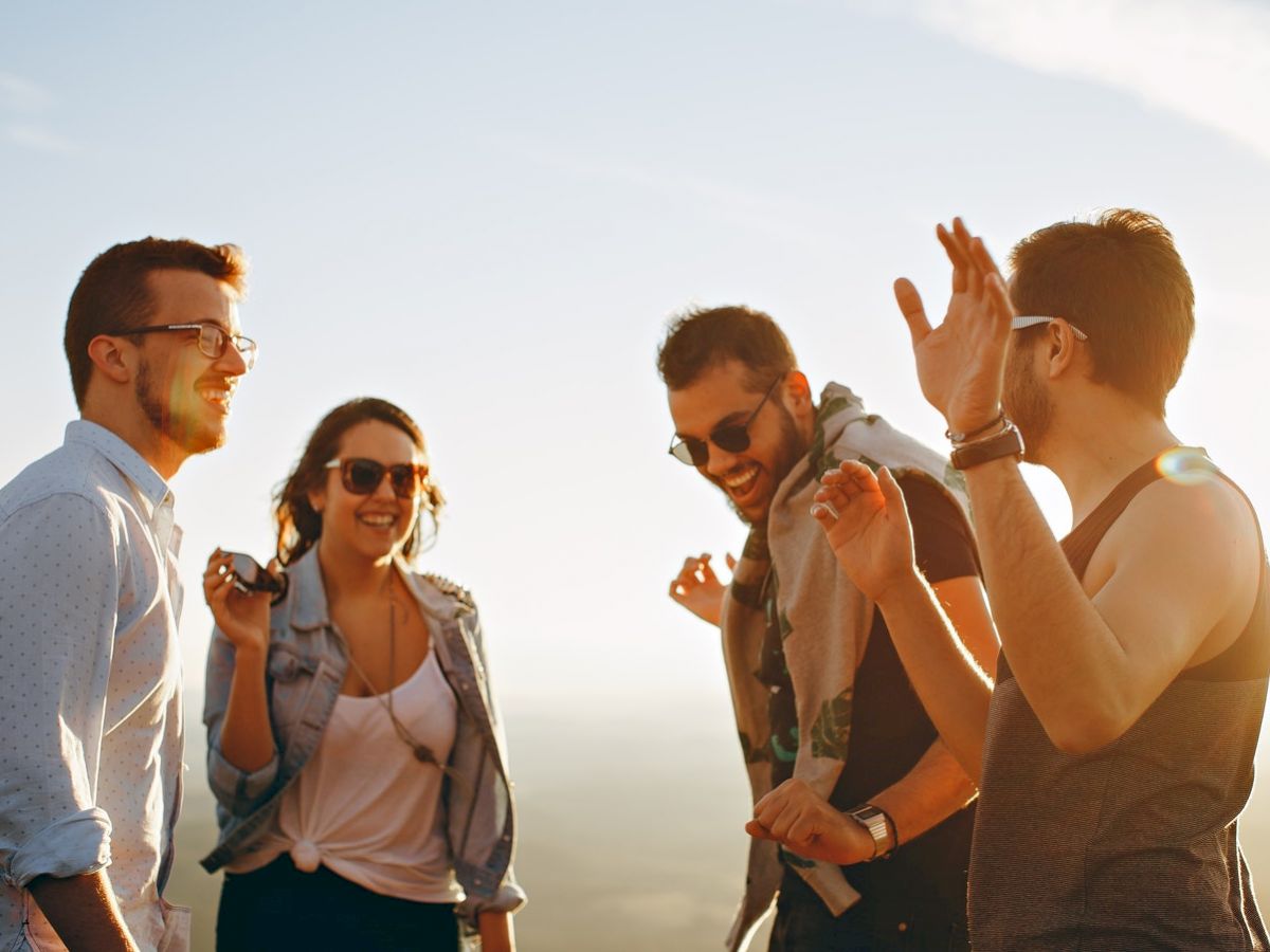 Four people are outdoors, laughing and enjoying themselves, with a bright sky in the background.