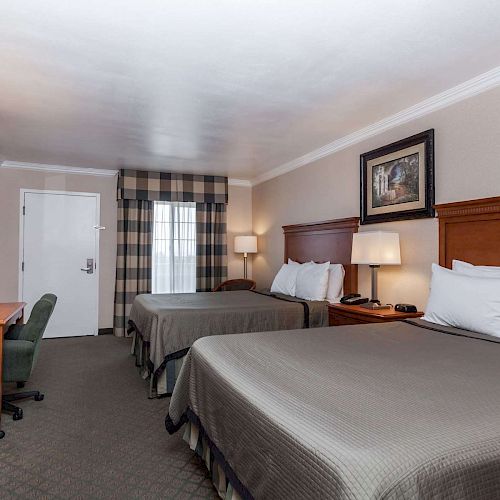 This image shows a hotel room with two double beds, a TV, desk and chair, lamps, and a window covered by plaid curtains.