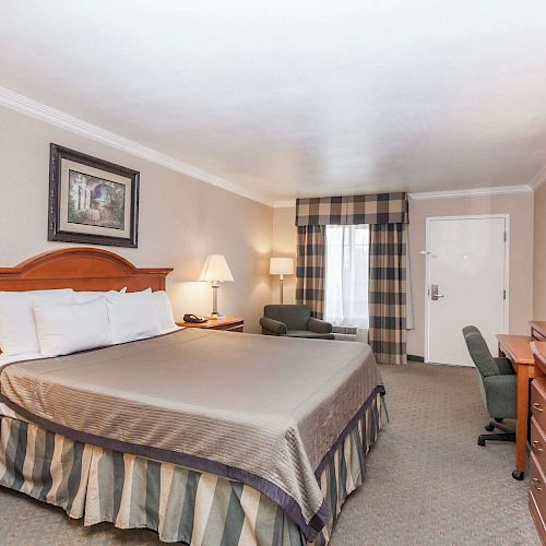 This image shows a hotel room with a large bed, two nightstands with lamps, a TV on a dresser, a desk with a chair, and a window with curtains.