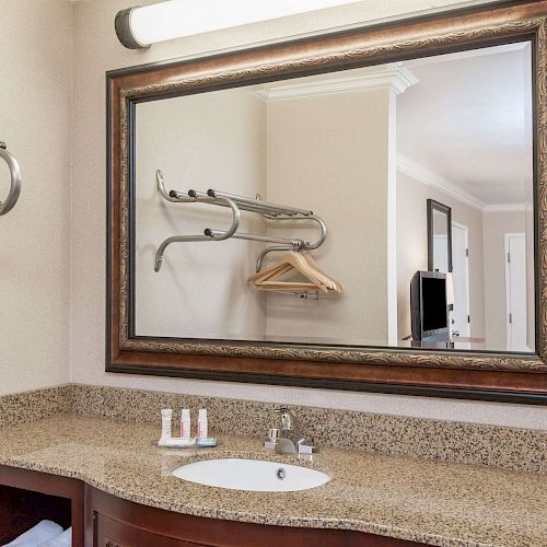 A bathroom vanity with a granite countertop, sink, and toiletries, a large framed mirror reflecting a hallway, and wall-mounted towel racks with hangers.