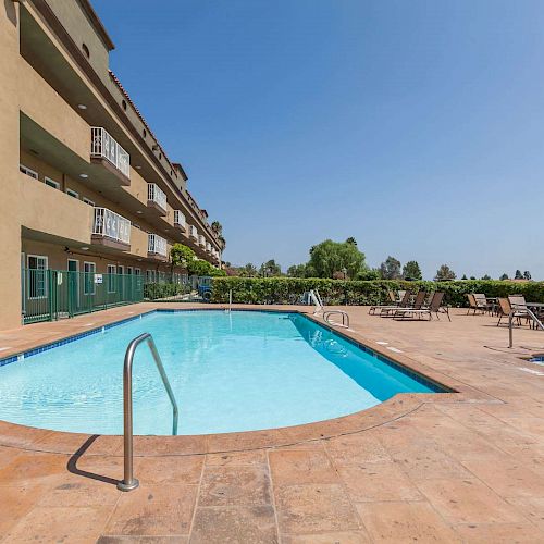 Image depicts an outdoor swimming pool and hot tub surrounded by a patio with lounge chairs near a multi-story building.