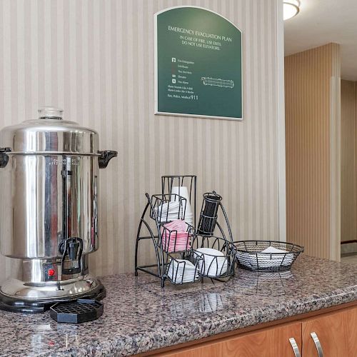 A coffee station with a large coffee urn, cups, lids, and condiments on a granite counter in a room with striped walls and a door in the background.