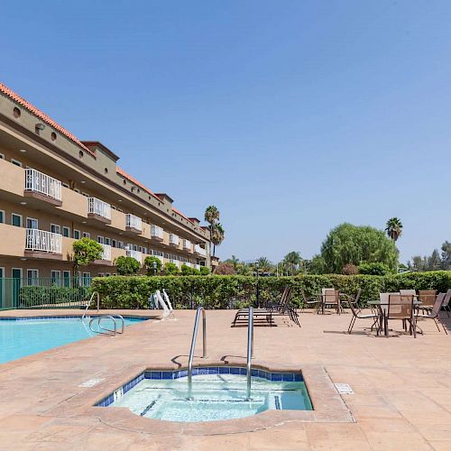 The image shows an outdoor pool area with a hot tub, surrounded by lounge chairs and tables, adjacent to a large, multi-story building.