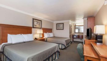 The image shows a hotel room with two double beds, a TV on a wooden desk, a lamp, and framed artwork on the walls.