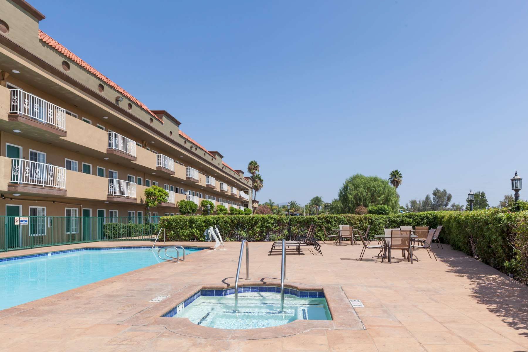 An outdoor pool area with a jacuzzi, deck chairs, and a multi-story building; clear skies and trees in the background.