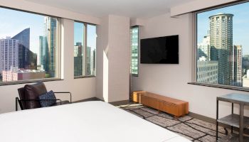 A modern hotel room with large windows showing a cityscape, a bed, a TV on the wall, a chair, and minimalistic furniture ends the sentence.