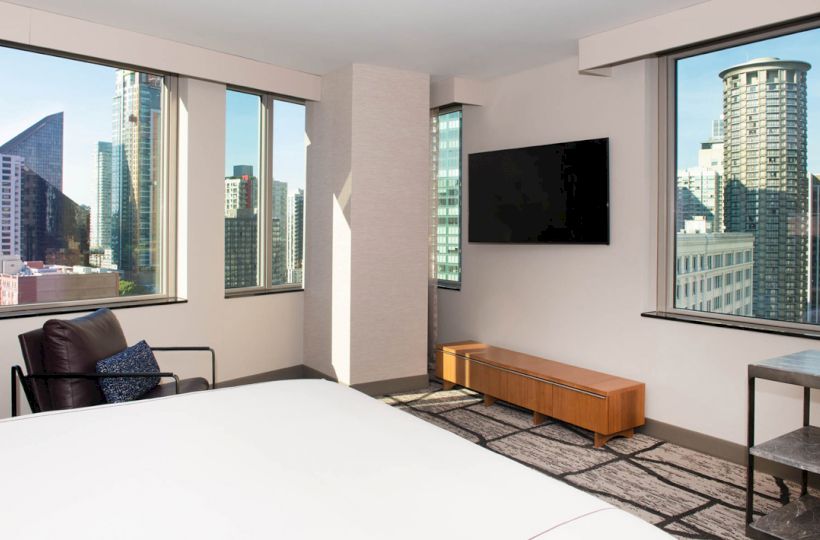 A modern hotel room with large windows showing a cityscape, a bed, a TV on the wall, a chair, and minimalistic furniture ends the sentence.