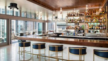The image shows a modern bar with a sleek design, featuring a long counter, blue bar stools, shelves stocked with various bottles, and large windows.