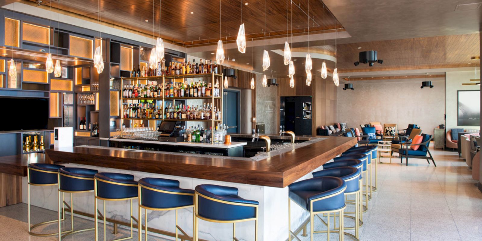 The image shows a modern bar with sleek design, featuring a well-stocked liquor display, a row of blue barstools, and pendant lights hanging above.