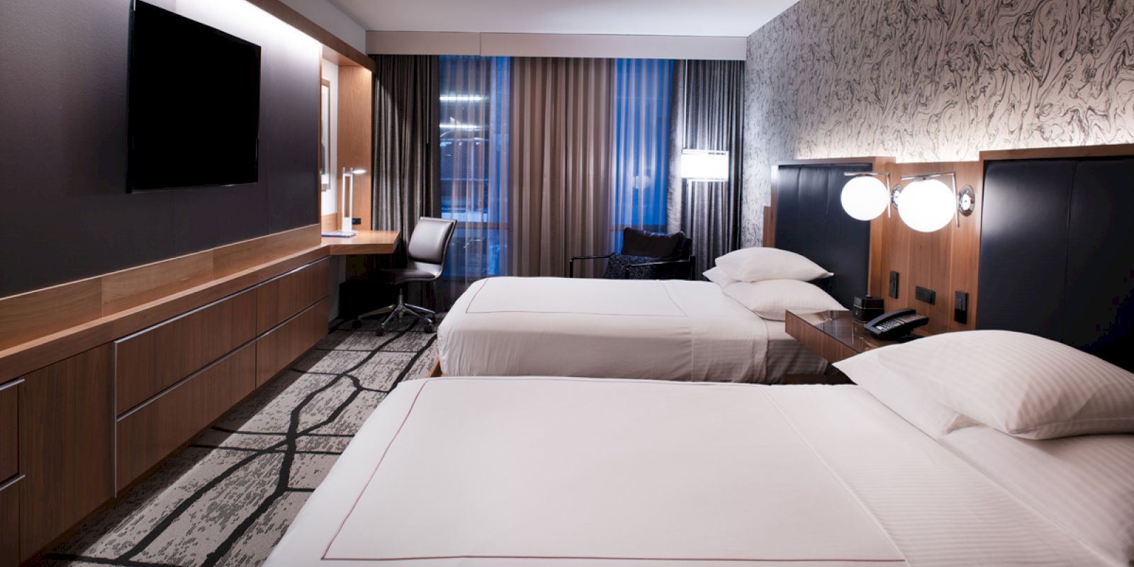 The image depicts a modern hotel room with two neatly made beds, a large television, a work desk and chair, and stylish decor.