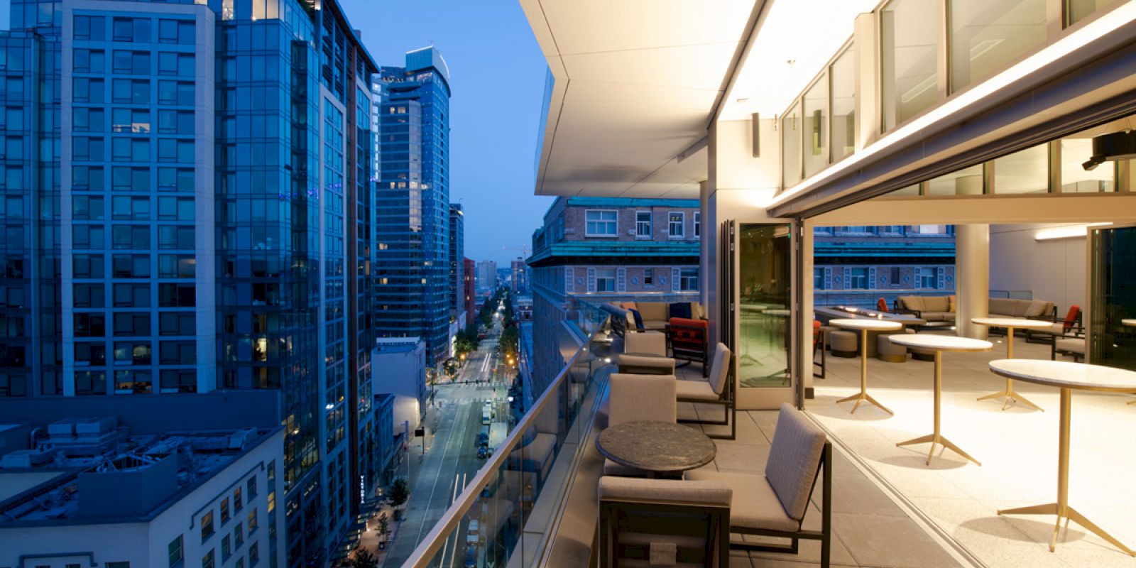 A modern rooftop terrace with tables and chairs overlooking a cityscape with tall buildings during twilight, city lights illuminating the scene.