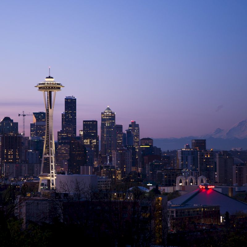A twilight view of a city skyline featuring a prominent tower, with a mountain visible in the background, likely Seattle's Space Needle and Mount Rainier.