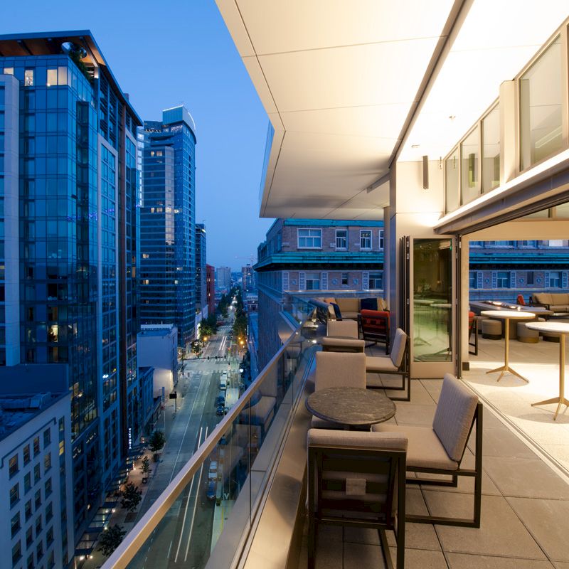 An outdoor balcony at night overlooks a cityscape with tall buildings, featuring tables, chairs, and modern lighting.