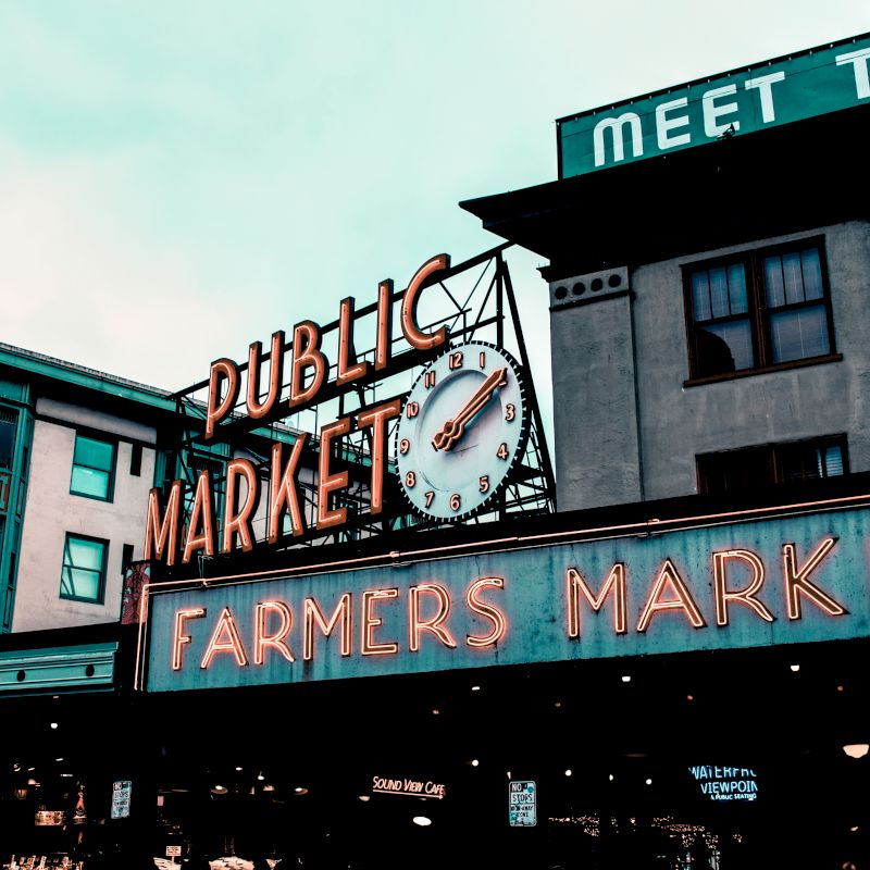 The image shows a historical building with neon signs for a "Public Market" and "Farmers Market," featuring a clock, likely indicating a bustling location.