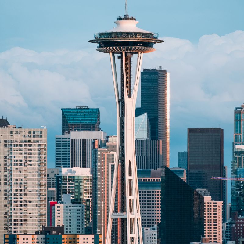 The image shows the Space Needle in Seattle, surrounded by modern skyscrapers and buildings with a background of cloudy skies.