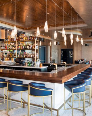 A modern bar with a wooden counter, blue barstools, and hanging lights. The bar is well-stocked, and there's seating in the back for patrons.
