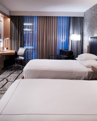 A modern hotel room with two beds, a wall-mounted TV, a desk, and contemporary decor featuring neutral tones and abstract patterns.