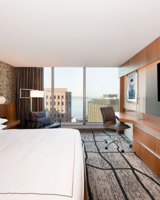 A modern hotel room with a large bed, a wall-mounted TV, a desk and chair, a window with city views, and contemporary decor.