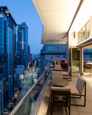 A modern cityscape at dusk with tall buildings and a well-furnished outdoor terrace featuring tables and chairs on the right.