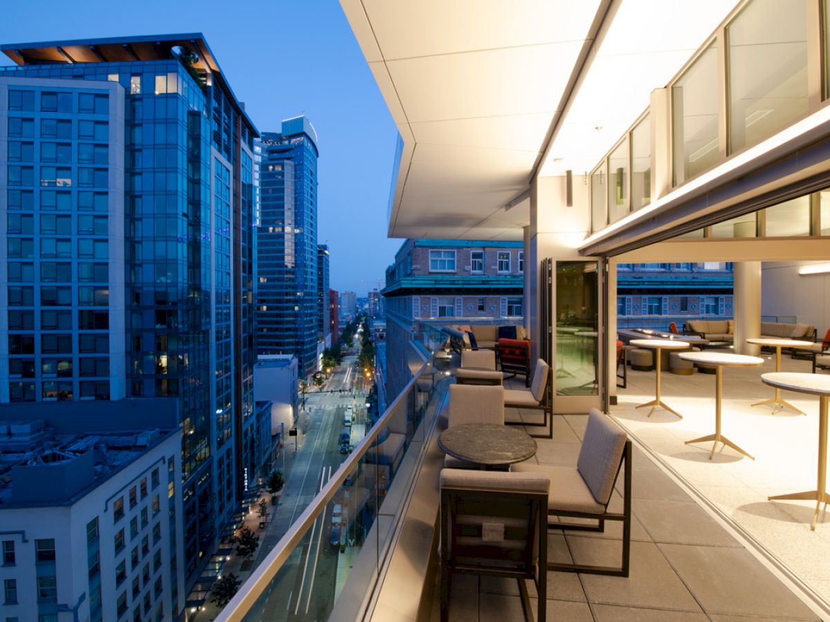 A modern rooftop patio overlooks a cityscape at dusk, featuring comfortable seating and tables, with tall buildings lining a street below.