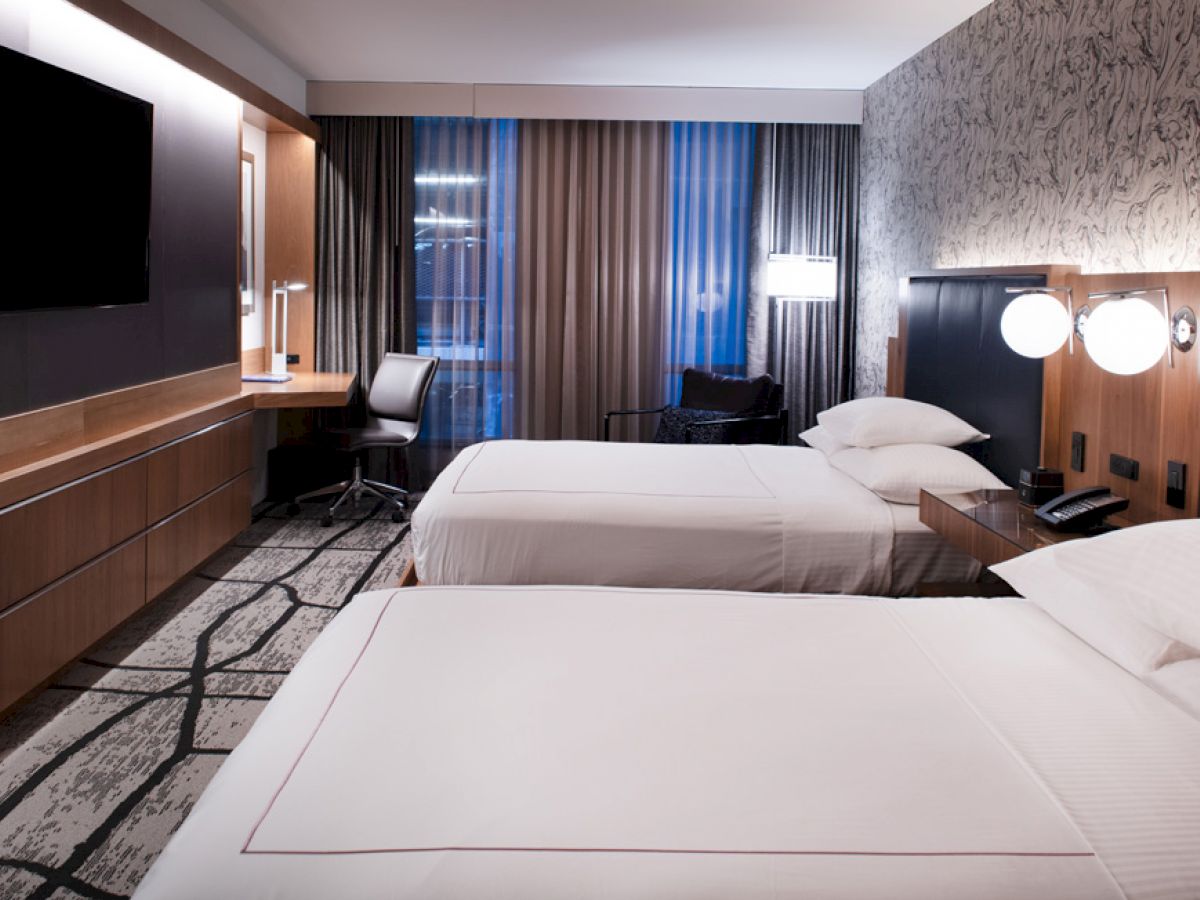 A modern hotel room features two neatly made beds, a wall-mounted TV, a work desk with a chair, lamps, and a patterned carpet.