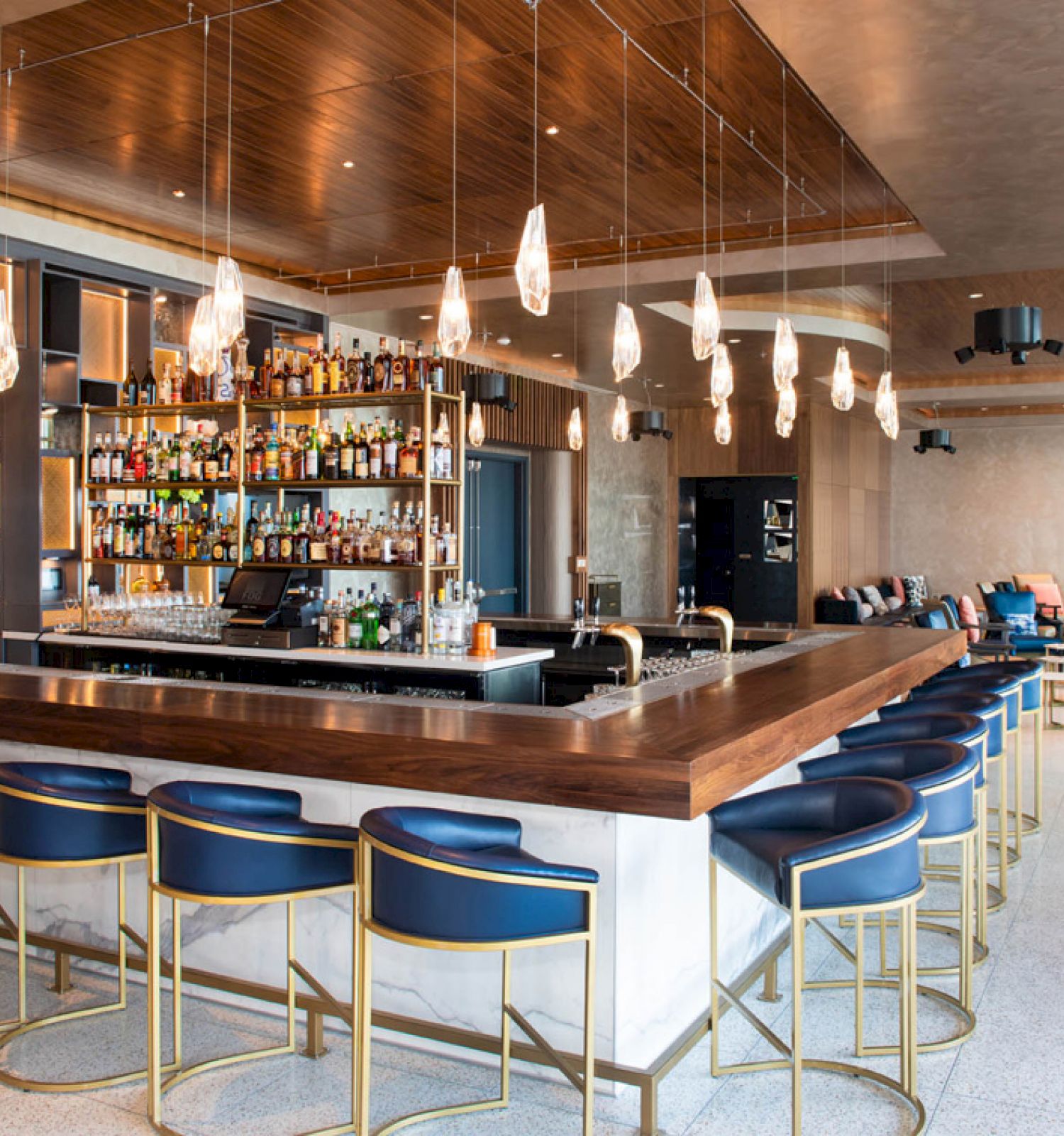 A modern bar with high stools, a variety of drinks on shelves, hanging pendant lights, and seating areas in the background.
