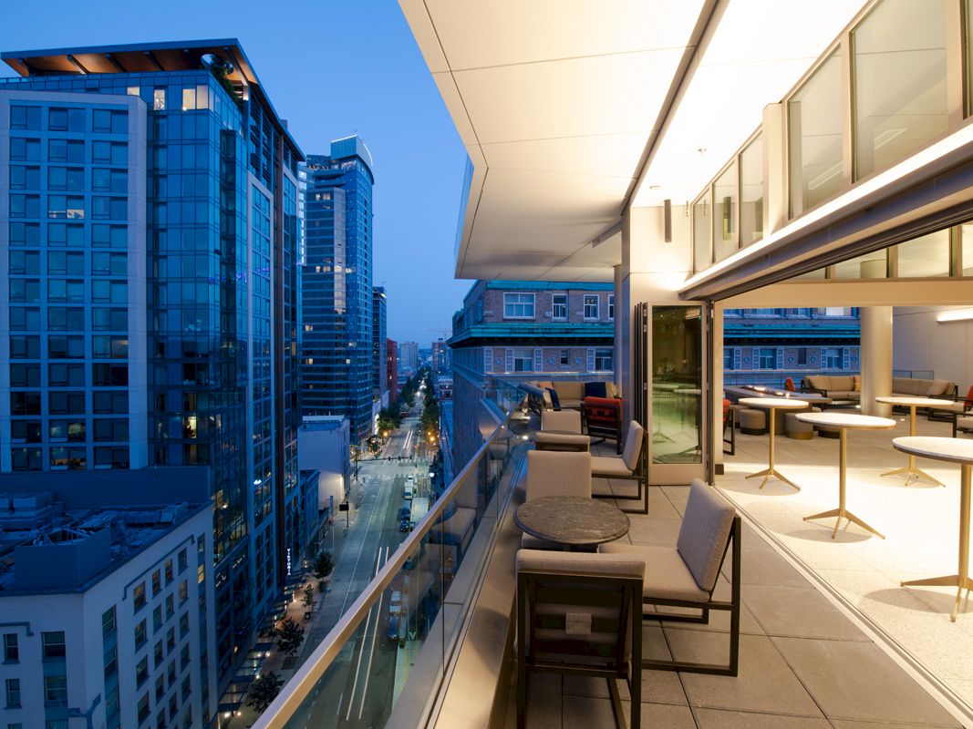 This image shows a modern outdoor balcony with seating, overlooking a city street lined with tall buildings during the evening.