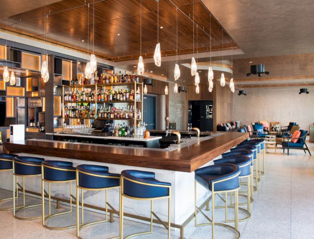 The image shows a modern, well-lit bar with a wooden countertop, blue cushioned chairs, hanging lights, and a stocked back shelf.