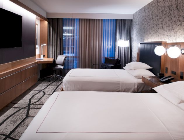 A modern hotel room with two neatly made beds, a flat-screen TV, a desk with a chair near the window, and stylish decor.