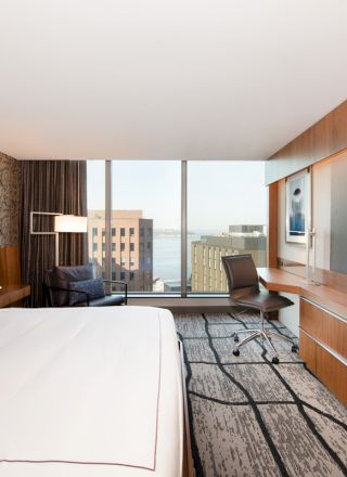 A modern hotel room with a large bed, a work desk and chair, a wall-mounted TV, and a window with a view of buildings.