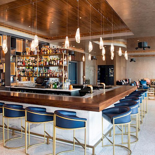 The image shows a modern bar with a long counter, several bar stools, hanging lights, and a well-stocked back bar, all in a sleek interior design.