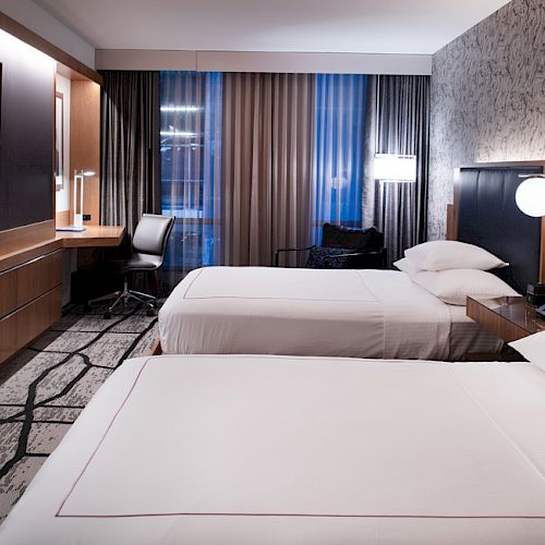 A modern hotel room features two neatly made beds, a wall-mounted TV, a work desk with a chair, and stylish decor including textured wallpaper and lighting.