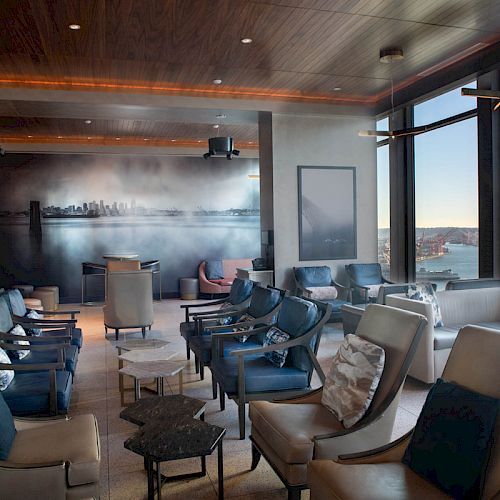 A modern lounge with multiple chairs and tables, large windows offering a scenic waterfront view, and wall art depicting a cityscape.
