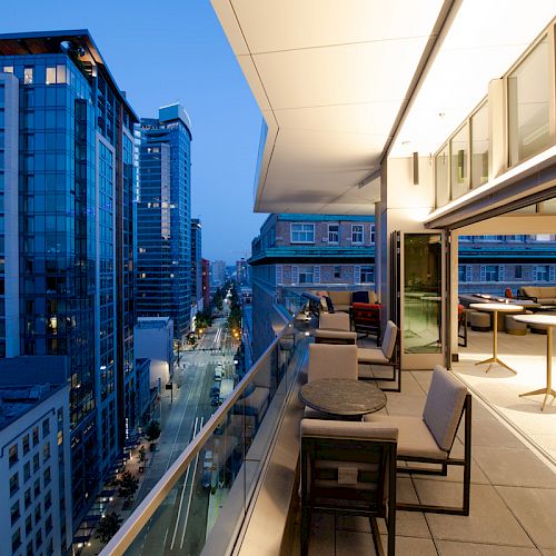 An outdoor rooftop terrace overlooking a cityscape at dusk, with modern furniture and a view of surrounding tall buildings lining the street.