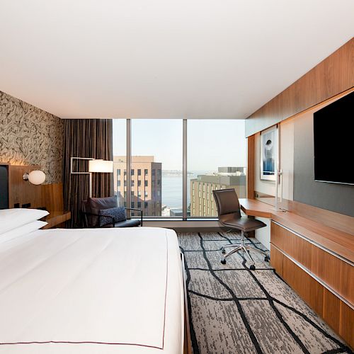 This image shows a modern hotel room with a large bed, a desk area, a flat-screen TV, and a city view through a large window.