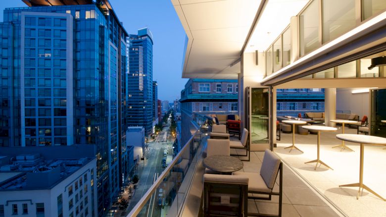 A modern cityscape at dusk, with high-rise buildings. The image features an outdoor patio with seating, tables, and an illuminated indoor area.