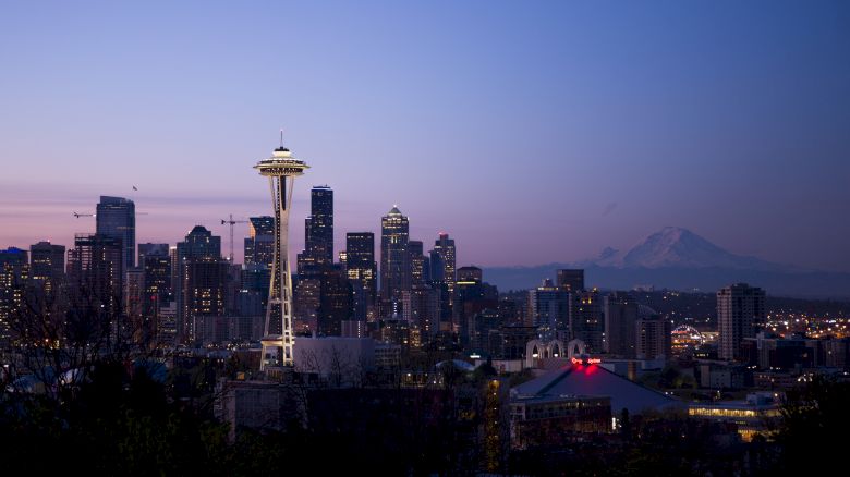 This image shows the skyline of Seattle, including the iconic Space Needle, against a backdrop of a mountain at dusk with a clear, colorful sky.