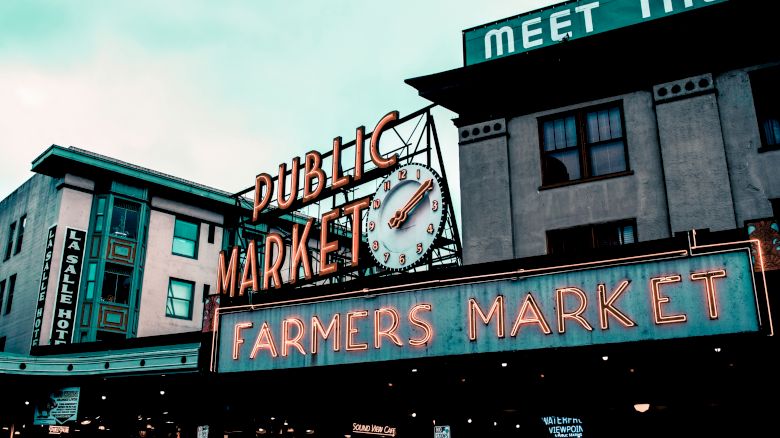 The image shows a neon sign for a Public Market/Farmers Market in an urban setting, with buildings and a clock in the background.