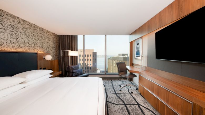 A modern hotel room with a large bed, wall-mounted TV, desk, and floor-to-ceiling window offering a city view.