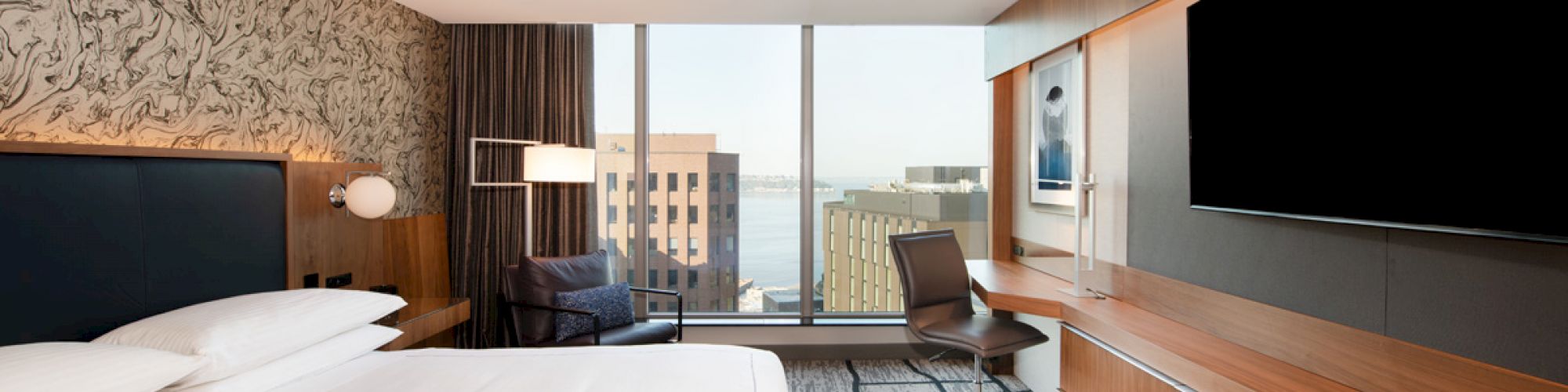 A modern hotel room features a large bed, wall-mounted TV, desk, chair, and window with a cityscape view.