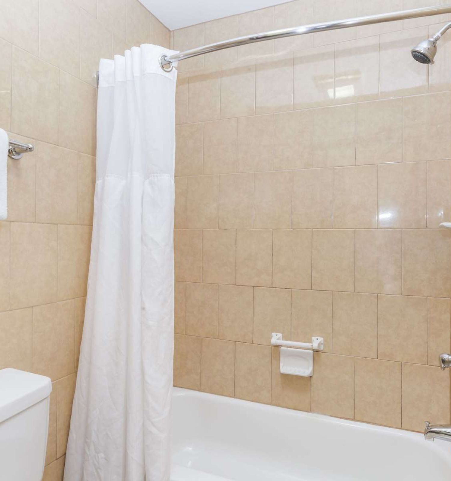 A clean bathroom with tan tile walls features a bathtub/shower combo, a white shower curtain, a towel hanging above the toilet, and a wall-mounted showerhead.