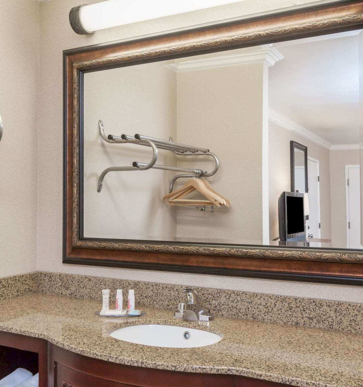 The image shows a bathroom vanity with a granite countertop, a sink, several small toiletries, a large mirror, and a rack with hangers.