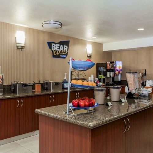 This image shows a breakfast buffet area with various food items and beverages on a countertop, including fruits and dispensers.