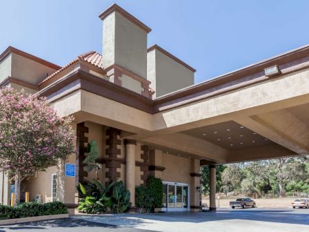 The image shows the entrance of a beige building with multiple chimneys, likely a hotel, featuring a covered driveway and a tree.