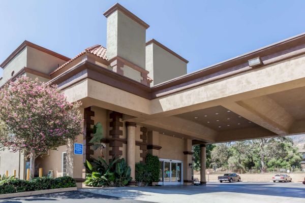 The image shows the entrance of a beige building with multiple chimneys, likely a hotel, featuring a covered driveway and a tree.