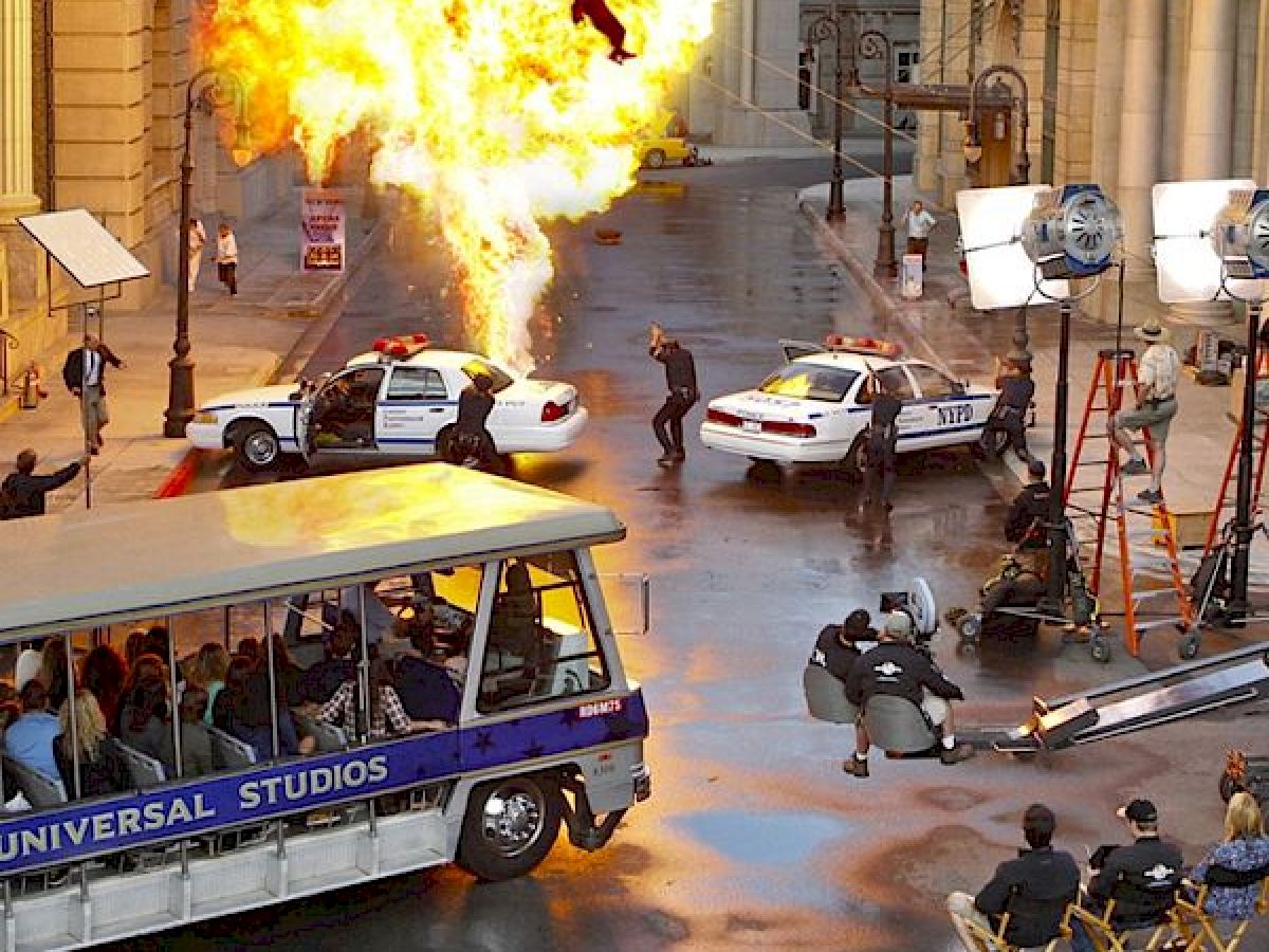 A movie scene is being filmed with a large explosion, police cars, and a film crew, while a Universal Studios bus tour passes by in the foreground.