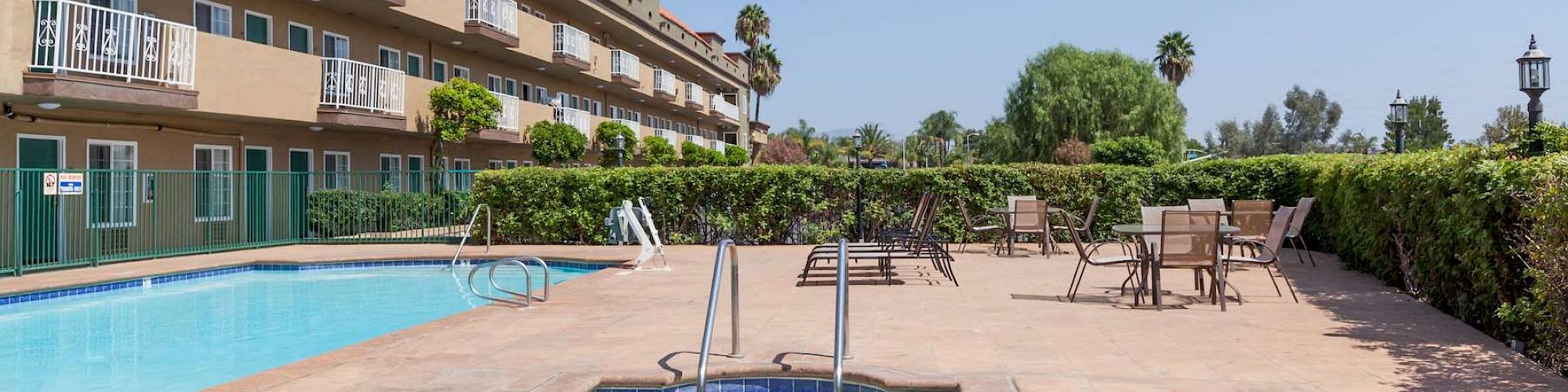 The image shows a pool area with a hot tub and some poolside seating. A multi-story building with balconies lines one side. It's a sunny day.